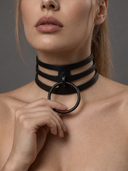 A close-up of a woman wearing a black multi-strand leather collar choker with a large circular metal ring at the front, set against a neutral gray background.
